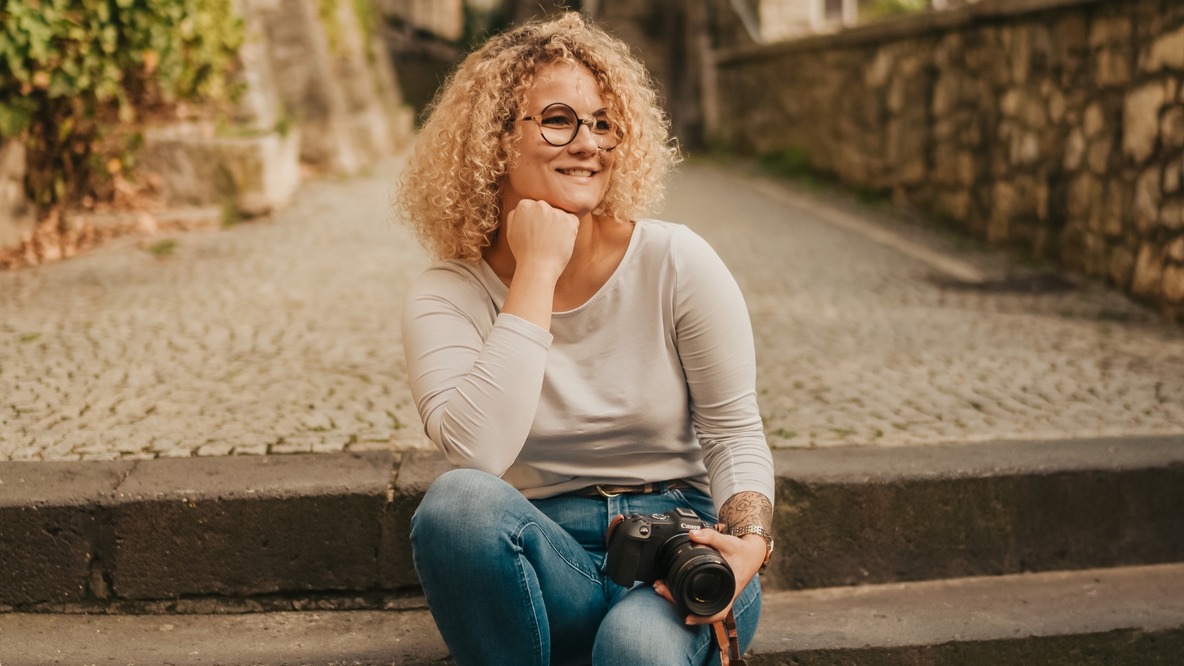 Fröhliche Fotografin mit lockigem Haar und Brille sitzt auf Treppe mit Kamera.